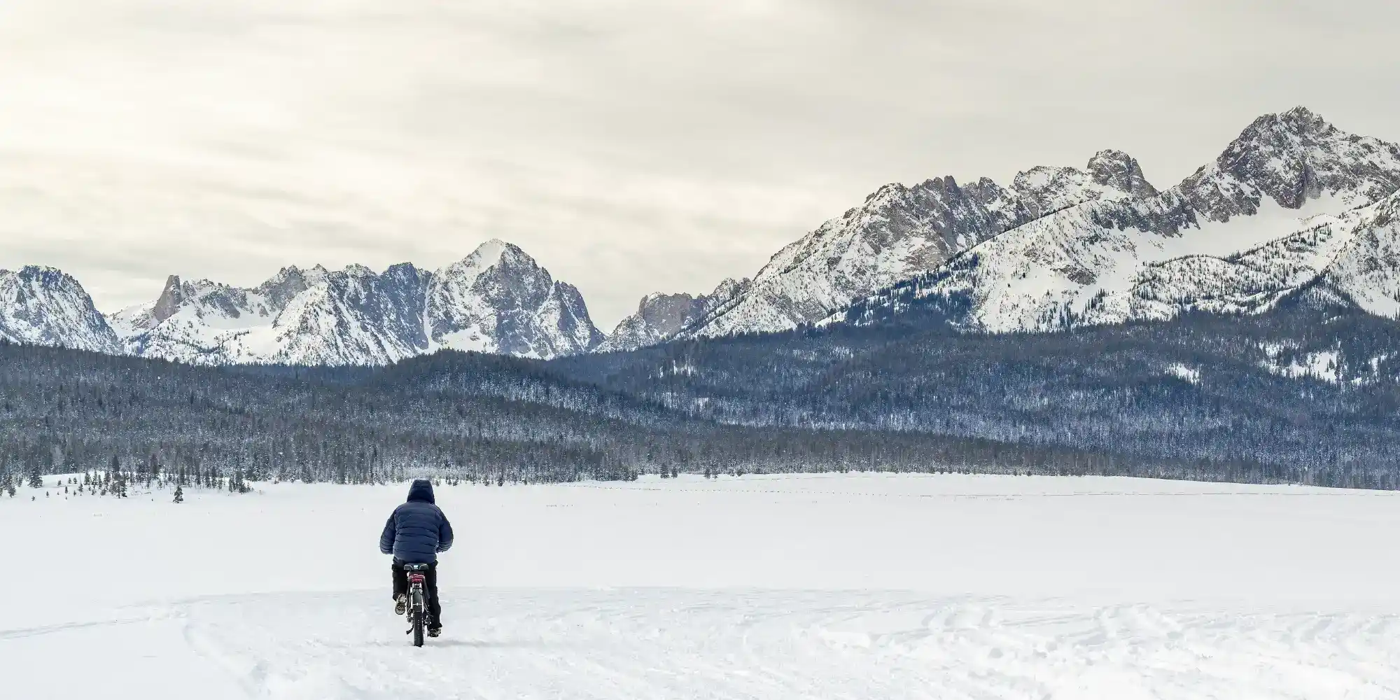 on a snowy plain with snowcovered mountains in the background an ebike rider travels down a well-travelled snow track